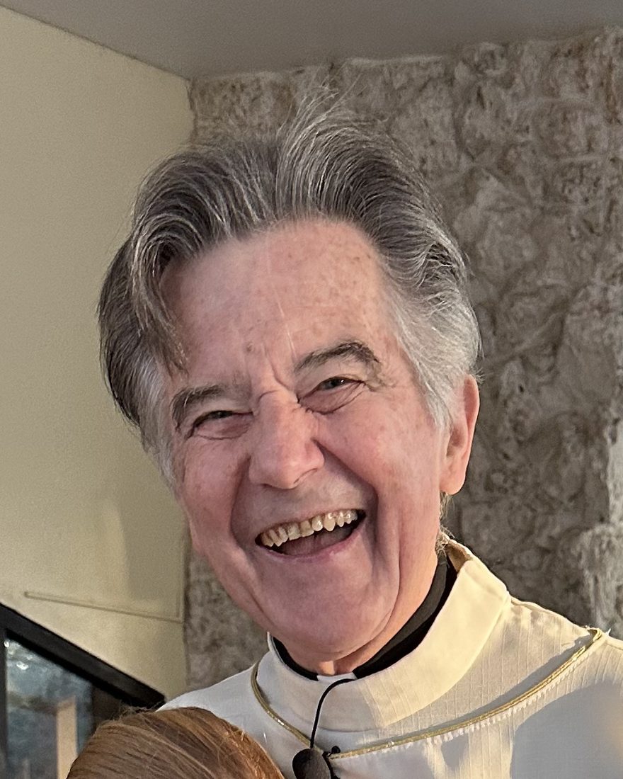 Fr. Danny Murphy laughing, in a white alb with a green cross, outside Holy Cross Church.
